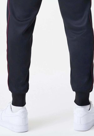 Untere Beine in schwarzen Hosen mit roten Seitenstreifen und weißen Nike-Sneakern mit sichtbaren Logos, stehend auf einer hellen Oberfläche.