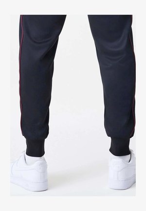 Jambes inférieures portant un pantalon noir avec des bandes rouges sur les côtés et des baskets blanches Nike avec des logos visibles, debout sur une surface claire.