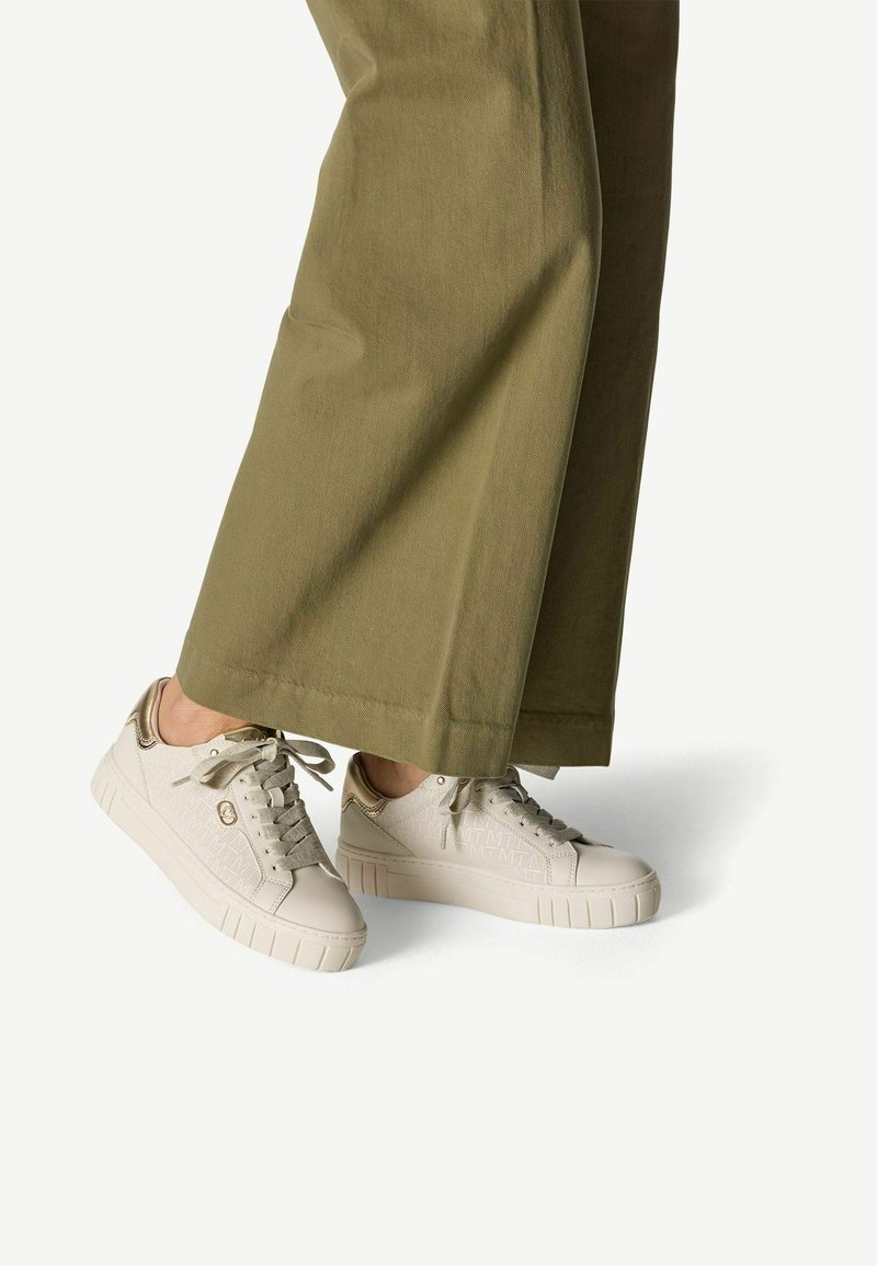 White sneakers with a textured surface and gold accents, featuring a flat sole and lace closure, paired with olive green wide-leg pants.