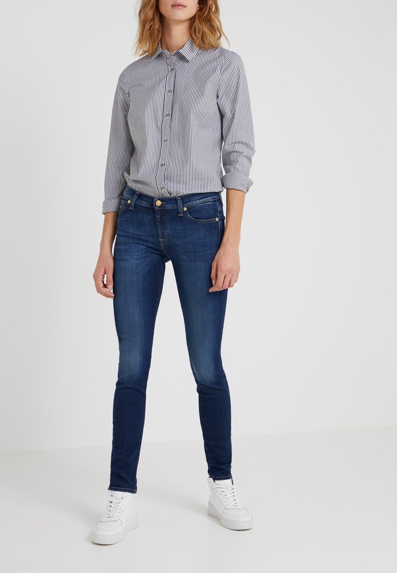 Randig skjorta i ljusblått och vitt, tillsammans med mörkblå slim-fit jeans och vita sneakers, stående mot en enfärgad bakgrund.