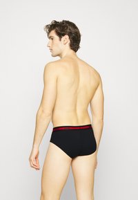 Bóxers para hombres negros con una cinturilla elástica roja que lleva la marca "HUGO". Tejido suave y ajuste ceñido, visto desde la parte posterior.