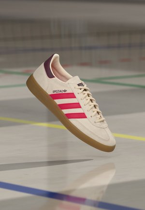 Sneakers Adidas Spezial blanc cassé avec des bandes rouges, semelle en gomme et accent bordeaux au talon, montrées en suspension au-dessus du sol d’un terrain de sport.