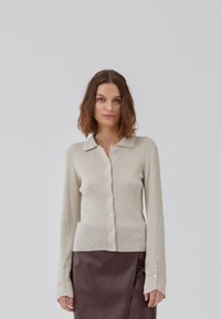 Cardigan beige en tricot côtelé avec un col, des manches longues et des boutons sur le devant. Associé à une jupe noire élégante.