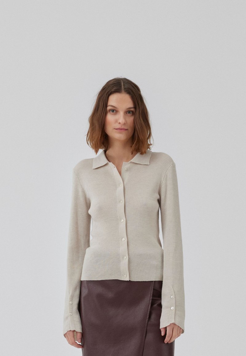 Cardigan beige en tricot côtelé avec un col, des manches longues et des boutons sur le devant. Associé à une jupe noire élégante.