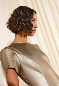 Metallik pronksirikas kleit siledast kangast, ümarate kaelustega ja puff-sügisestega lühikeste varrukatega, vaadatuna küljelt pehmete voltitud kangaste taustal.