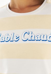 Randig kortärmad t-shirt med växlande vita och ljusbeige ränder, med blå text som säger "Table Chaud" i ett lekfullt teckensnitt.