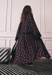 Kvinde med langt krøllet hår iført en sort kappe med røde og grønne læbestiftsaftryk, stående barfodet på et stribet tæppe nær en stribet fodskammel.