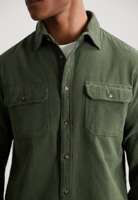 Green corduroy shirt with button-down collar, two chest pockets, and brown buttons. Fabric features a ribbed texture.