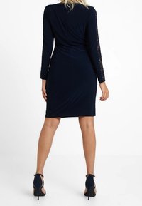 Marinblå klänning med långa ärmar, med spetsdetaljer på ärmarna, figurnära siluett och knälång nederkant, tillsammans med högklackade skor.