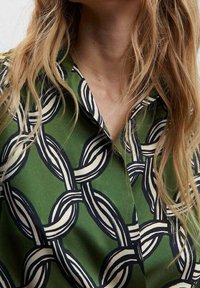 Grüne Bluse mit einem Kettenmuster in Schwarz und Weiß, mit Kragen und weicher Textur. Nahaufnahme der Stoffdetails.