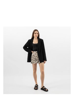 Giovane donna in piedi che indossa una giacca nera, un top nero, una gonna con stampa zebra e sandali neri su sfondo bianco uniforme.