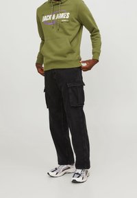 Felpa verde oliva con logo bianco e viola, abbinata a pantaloni cargo neri con tasche laterali. Le scarpe sono bianche con dettagli neri e rossi.