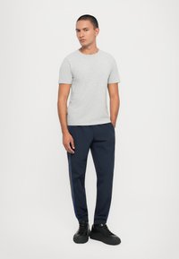 Pelēka īsām piedurknēm T-kreklā kombinēta ar tumši ziliem sporta biksēm ar sānu svītru. Modelis valkā melnus sporta apavus, radot ikdienišķu izskatu.