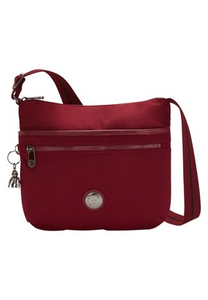 Petit sac bandoulière rouge avec poche zippée à l'avant, sangle réglable et logo de marque rond en métal à l'avant.