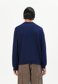 Hanorac de culoare bleumarin cu mâneci lungi, manșete ribitate și un tiv drept, confecționat dintr-un material moale. Pantaloni cargo maro vizibili la talie.