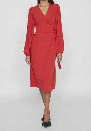Woman wearing a red wrap midi dress with long puffed sleeves and black pointed heels with bows, standing against a plain background.