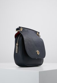 Granatowa skórzana torebka typu crossbody z klapką, złotymi akcentami i zamykaną na zamek boczną kieszenią. Teksturowana powierzchnia z gładką podszewką.