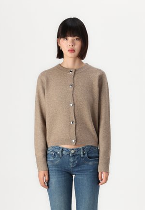 Beige bolero laget av mykt strikkemateriale, med rund hals og sølv hjerteformede knapper foran. Kombinert med blå jeans.