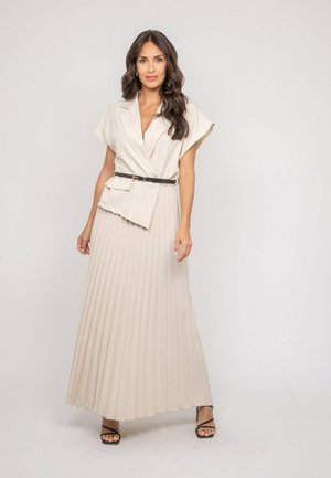 Femme aux longs cheveux bruns portant une robe maxi plissée beige avec une ceinture noire et des talons noirs à brides, debout devant un fond uni.