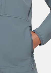 Giacca softshell grigia con tasca con zip, texture liscia e polsini aderenti. Il tessuto è impermeabile e flessibile per un uso attivo.