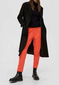 Schwarzer langer Mantel über einem schwarzen Rollkragenpullover, kombiniert mit leuchtend orangefarbenen tapered Hosen und schwarzen Knöchelstiefeln mit einer robusten Sohle.