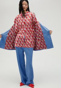Camicia button-up con un motivo geometrico in tonalità rosse e rosa, abbinata a pantaloni blu e scarpe rosse a punta.