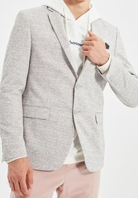 Blazer grigio chiaro con texture, due bottoni, rever a punta e tasche laterali, indossato sopra un hoodie bianco. Leggero motivo a puntini su tutta la superficie.