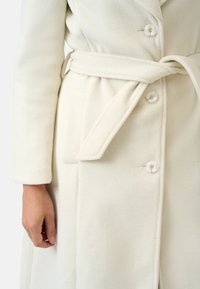 Cream-colored wool coat featuring a soft texture, a shawl collar, two large buttons, and a tied belt at the waist. Two side pockets visible.