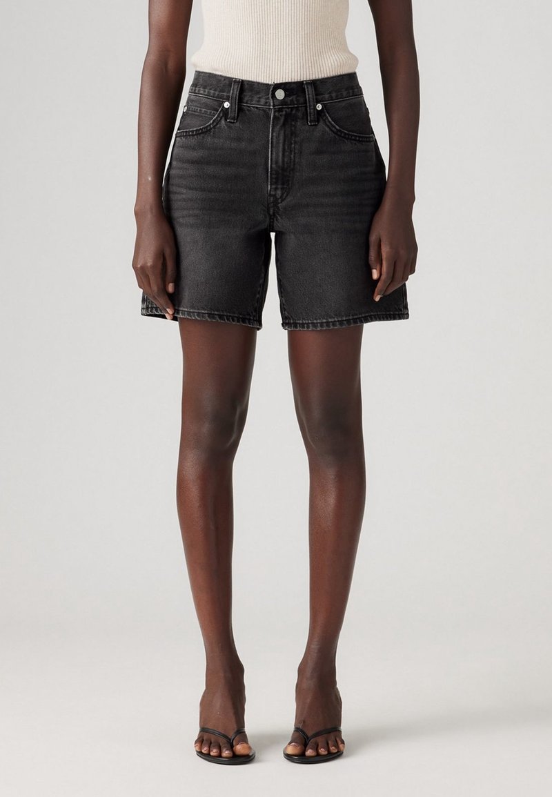 Personne portant un short en denim noir taille haute, un haut blanc sans manches et des sandales noires à bout ouvert, debout devant un fond uni.