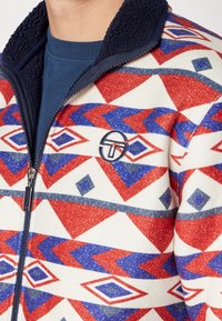Fleece ritsjack met een rood, wit en blauw geometrisch patroon. Het heeft een marineblauwe kraag, een zachte textuur en een logo appliqué op de borst.