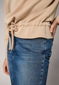 Beige långärmad topp med dragsko i midjan, tillsammans med blå denimjeans; handen vilar i jeansfickan.