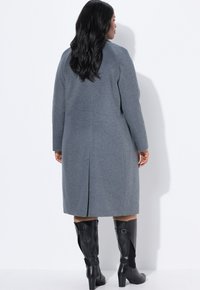 Cappotto grigio in misto lana con maniche lunghe e spacco posteriore, visto da dietro, abbinato a stivali neri in pelle con tacco alti fino al ginocchio.