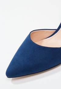 Chaussure à talon haut en daim bleu marine à bout pointu avec doublure intérieure en cuir beige sur fond blanc.