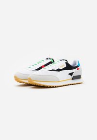 Paire de baskets blanches Puma avec des touches noires, bleues, rouges et vertes, semelle jaune et grip texturé sur fond blanc.