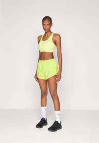 Conjunto de sujetador deportivo y pantalones cortos en verde neón, hecho de una tela ligera. Cuenta con un escote en forma de barco y una cinturilla elástica. Se acompaña de zapatillas negras.