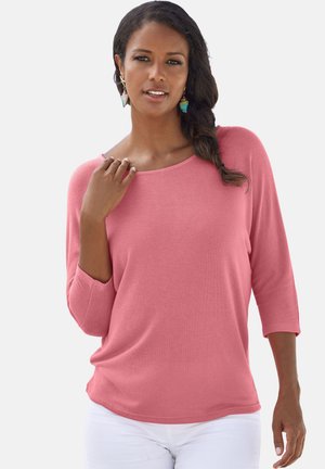 Rosa Langarmshirt mit weitem Halsausschnitt und lockerer Passform. Hergestellt aus einem weichen, leichten Stoff. Das Modell trägt weiße Hosen.