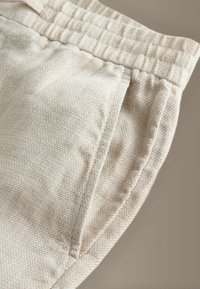 Gros plan sur un pantalon beige en tissu texturé avec une taille élastique et une poche latérale visible sur un fond neutre.