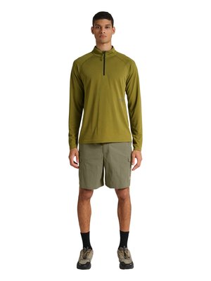 Jonge man staat frontaal, dragen een olijfgroene lange ritspullover, lichtolijfgroene cargoshorts, zwarte sokken en beige wandelschoenen.