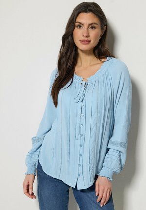 Kobieta o długich ciemnych włosach, ubrana w jasnoniebieską bluzkę na guziki z wiązaniem przy szyi i bufkami na rękawach, w połączeniu z niebieskimi dżinsami.