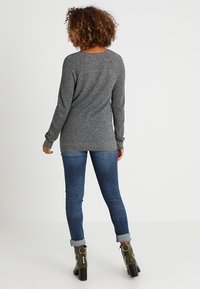 Maglione grigio a maniche lunghe con un motivo testurizzato, abbinato a jeans attillati blu scuro. La modella indossa stivaletti verde oliva con tacco spesso.