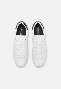 Fehér sneaker, sima bőrből készült felsőrésszel, lapos fűzőkkel és kontrasztos fekete sarkú akcentussal. Tartalmaz egy logót a nyelvén és a talpbetétjén.