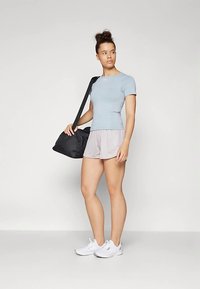 Svjetloplava sportska uska majica, sive kratke hlače, bijele tenisice, drži crnu torbu, stoji na neutralnoj pozadini.