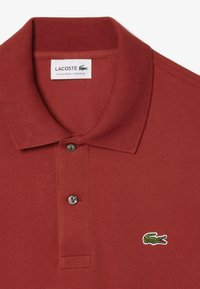 Röd polo-shirt i texturerat tyg, med kort krage, två knappar och en liten broderad grön krokodil-logotyp.