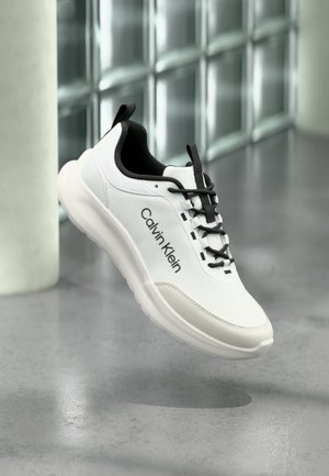 Bílá a černá teniska Calvin Klein s černými tkaničkami vznášející se nad šedou podlahou poblíž oken z skleněných bloků.