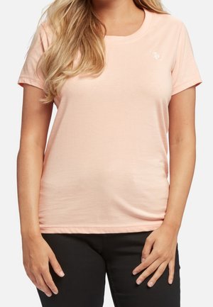 Nainen, jolla on vaalea persikanvärinen lyhythihainen t-paita, jossa on pieni valkoinen logo vasemmassa rinnassa, ja mustat housut, seisoo käsillä lähellä lantiota.