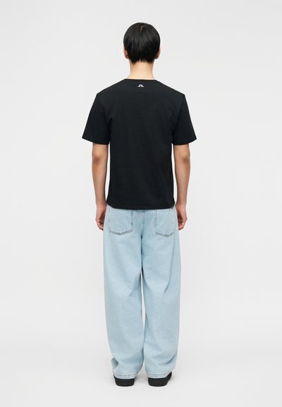 Άτομο που στέκεται με την πλάτη, φορά μαύρο T-shirt με μικρό λευκό λογότυπο στο πάνω μέρος της πλάτης και άνετο ανοιχτό μπλε τζιν.