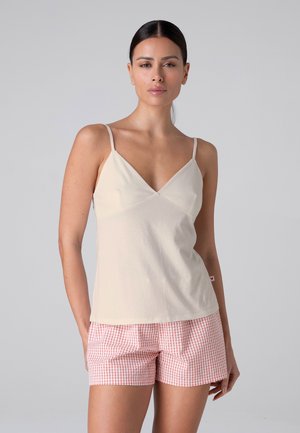 Donna con capelli scuri indossa un top crema con spalline sottili e pantaloncini rosa e bianchi a quadretti, in piedi davanti a uno sfondo grigio uniforme.