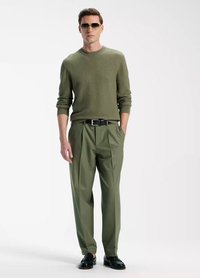 Olive green textured sweater, pleated olive trousers, and black belt. Model wears sunglasses and black shoes, standing against a neutral backdrop.