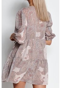 Kobieta z prostymi blond włosami, ubrana w luźną, sięgającą do kolan sukienkę w stonowany różowy i beżowy wzór paisley, widoczna z tyłu.