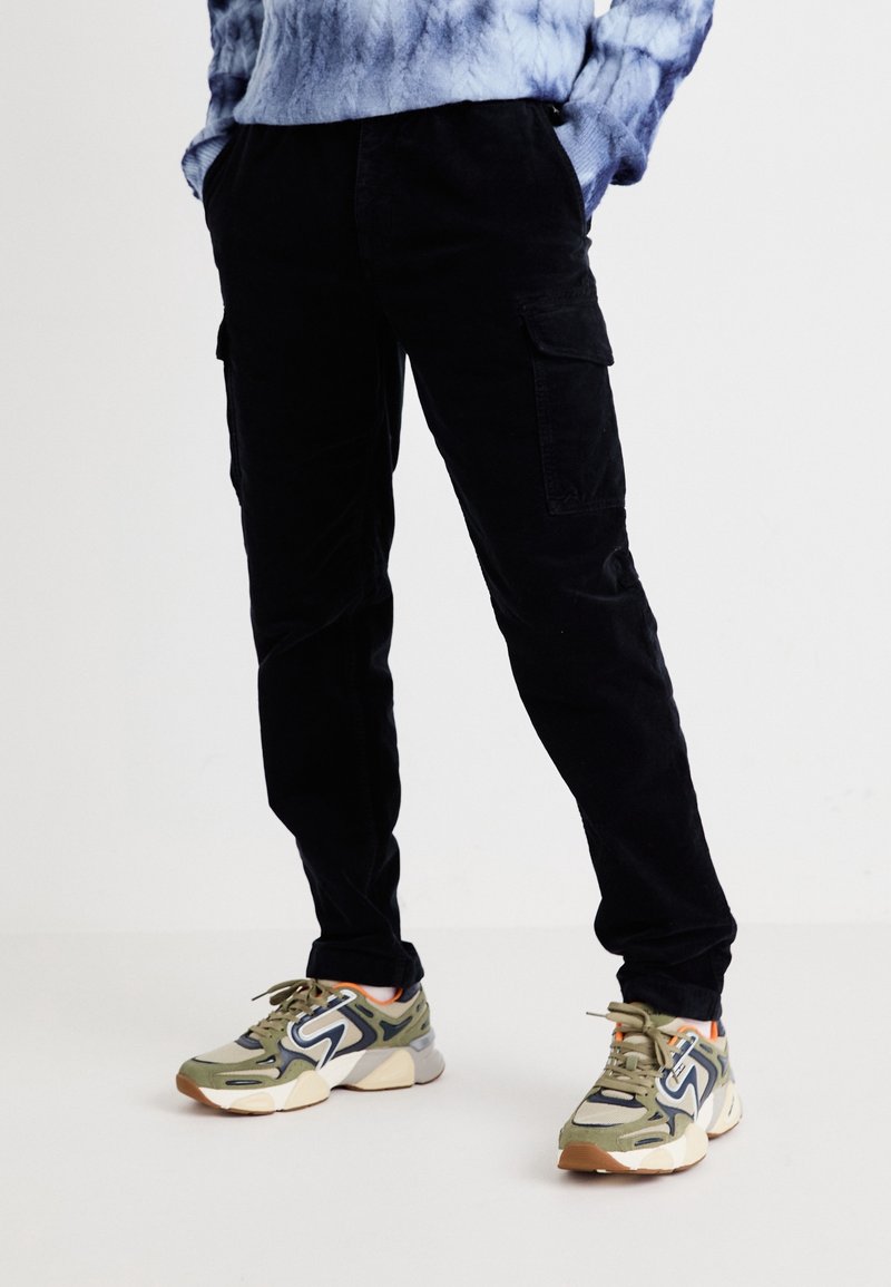 Personne portant un pantalon cargo noir et des baskets vertes, blanches et bleu marine, debout les mains dans les poches sur un fond blanc.